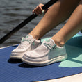 Load image into Gallery viewer, A girl sitting on a paddle board in the water wearing Camp Shoes. The shoes are a white color with teal accents.
