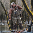 Load image into Gallery viewer, Man standing in water by a tree blowing a duck call