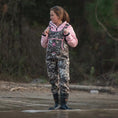 Load image into Gallery viewer, Woman walking by lake looking away while wearing Gator Waders Retro Waders in Seven camo with pink trim.