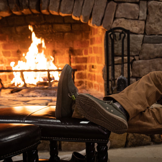 A guy propping his feet up by a fireplace while wearing a pair of olive Camp Shoes.