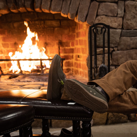 A guy propping his feet up by a fireplace while wearing a pair of olive Camp Shoes.