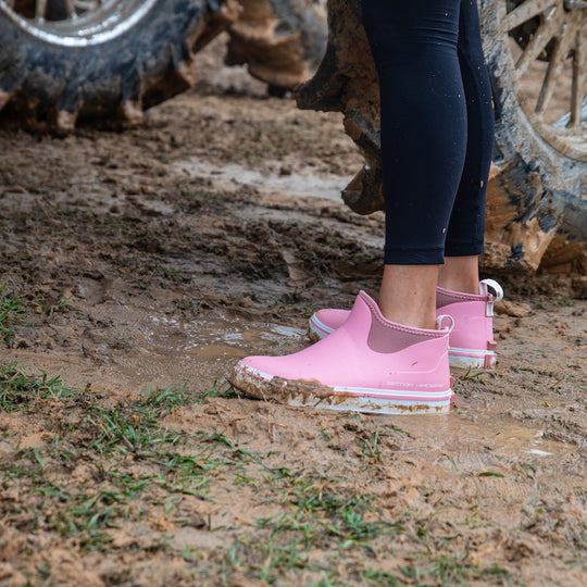 A pair of pink Air Mesh Camp Boots on a girls feet standing in the mud. 