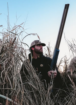 A man standing in a hunting blind wearing a black Gator Waders Highball Hoodie.