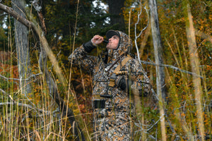 A guy standing in the woods blowing on a duck call wearing a pair of Gator Waders in 7 Brown camo. 