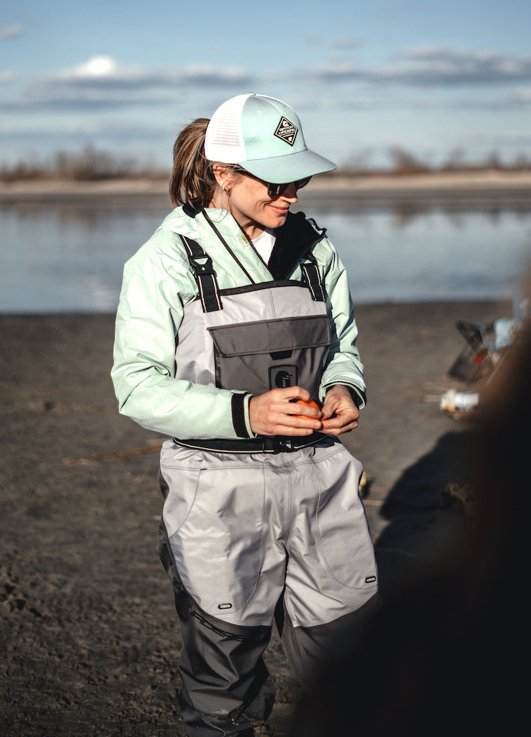 A girl standing by the water in a pair of Gator Waders grey Uninsulated Swamp Waders and a Sage color Bog Hoodie. 