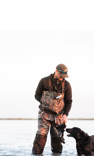 A guy standing in the water giving his dog a duck wearing a pair of Gator Waders Omega Zip Waders. 