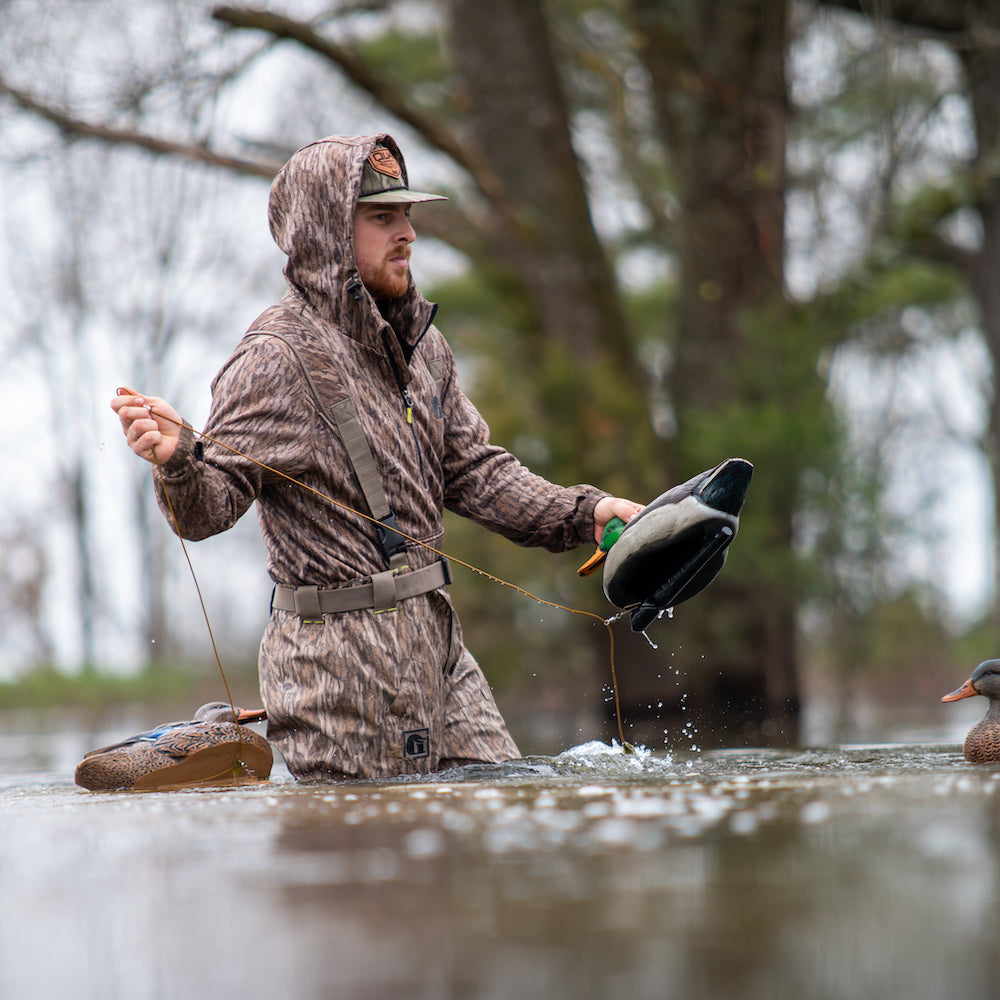 Man in camouflage waders and jacket holding a duck decoy in a flooded area with trees in the background.